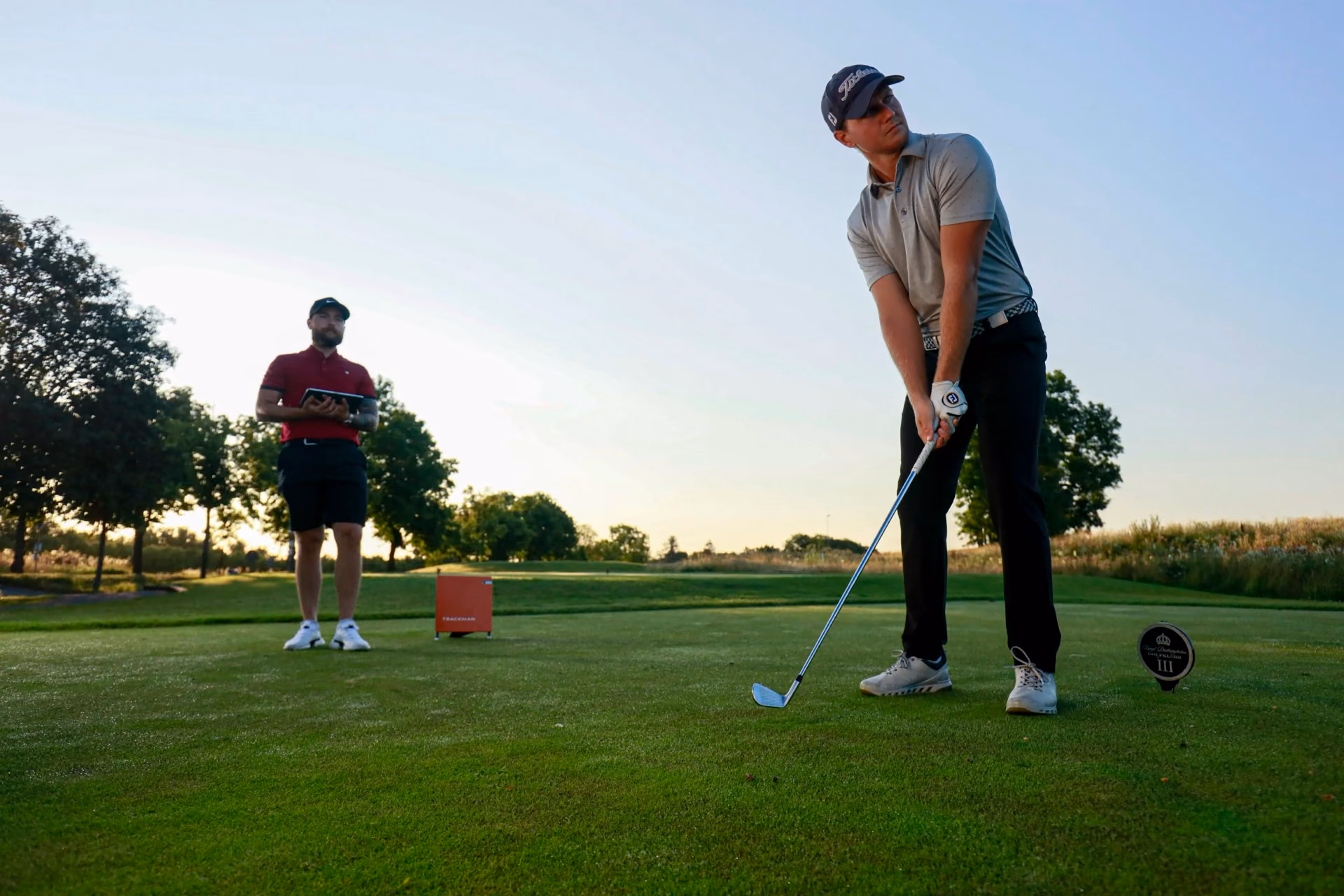 Un golfeur prépare un coup de fer sous le regard de son enseignant, tablette en main.