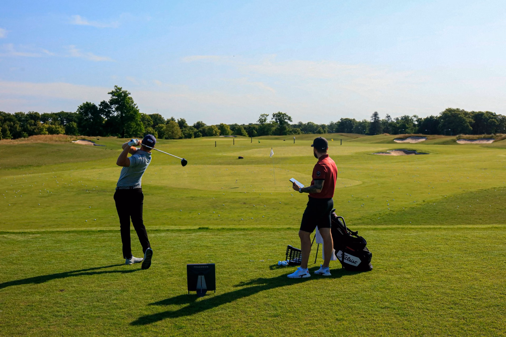 Un golfeur termine son swing au practice, avec un radar Trackman derrière lui et un enseignant tenant une tablette.