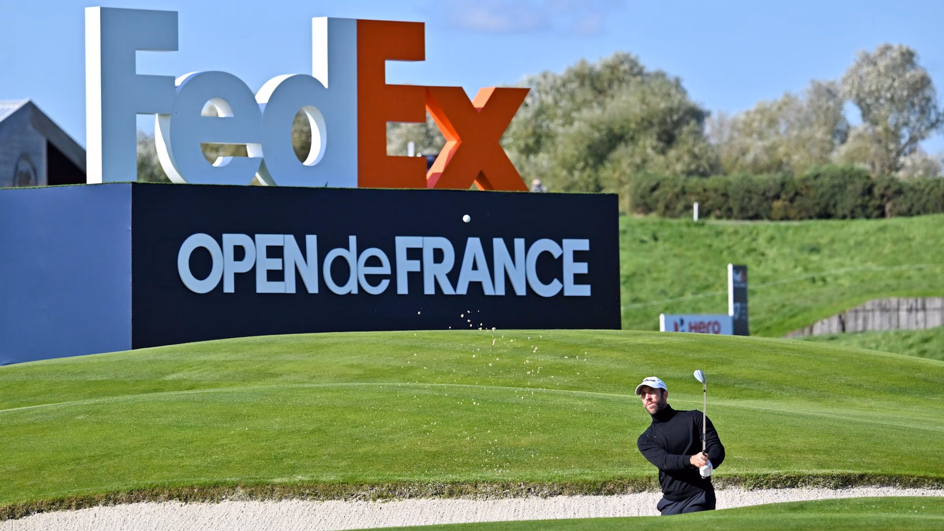 Un golfeur sort une balle du bunker devant le panneau FedEx Open de France.