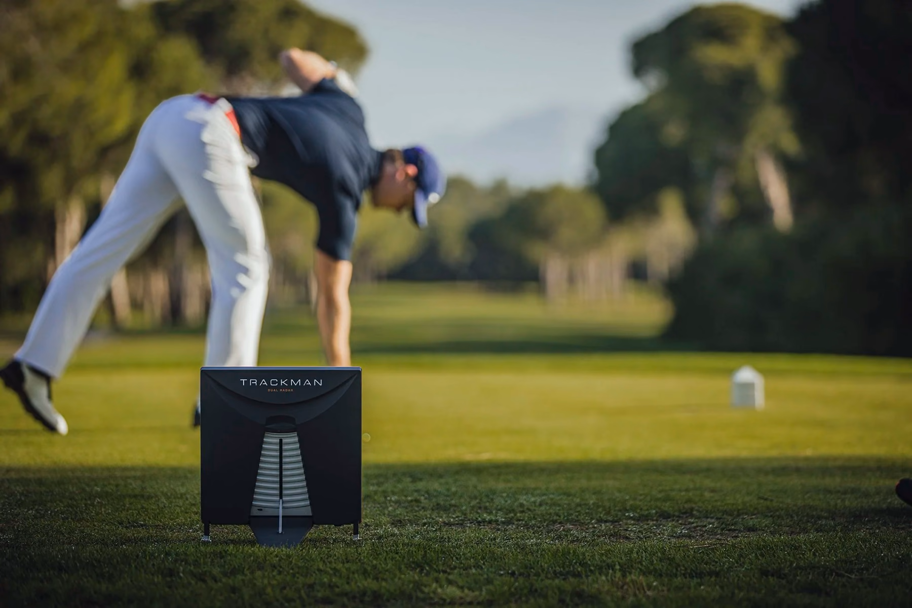 Un radar Trackman 4 installé derrière la zone de frappe, avec un golfeur qui prépare sa balle sur le tee.